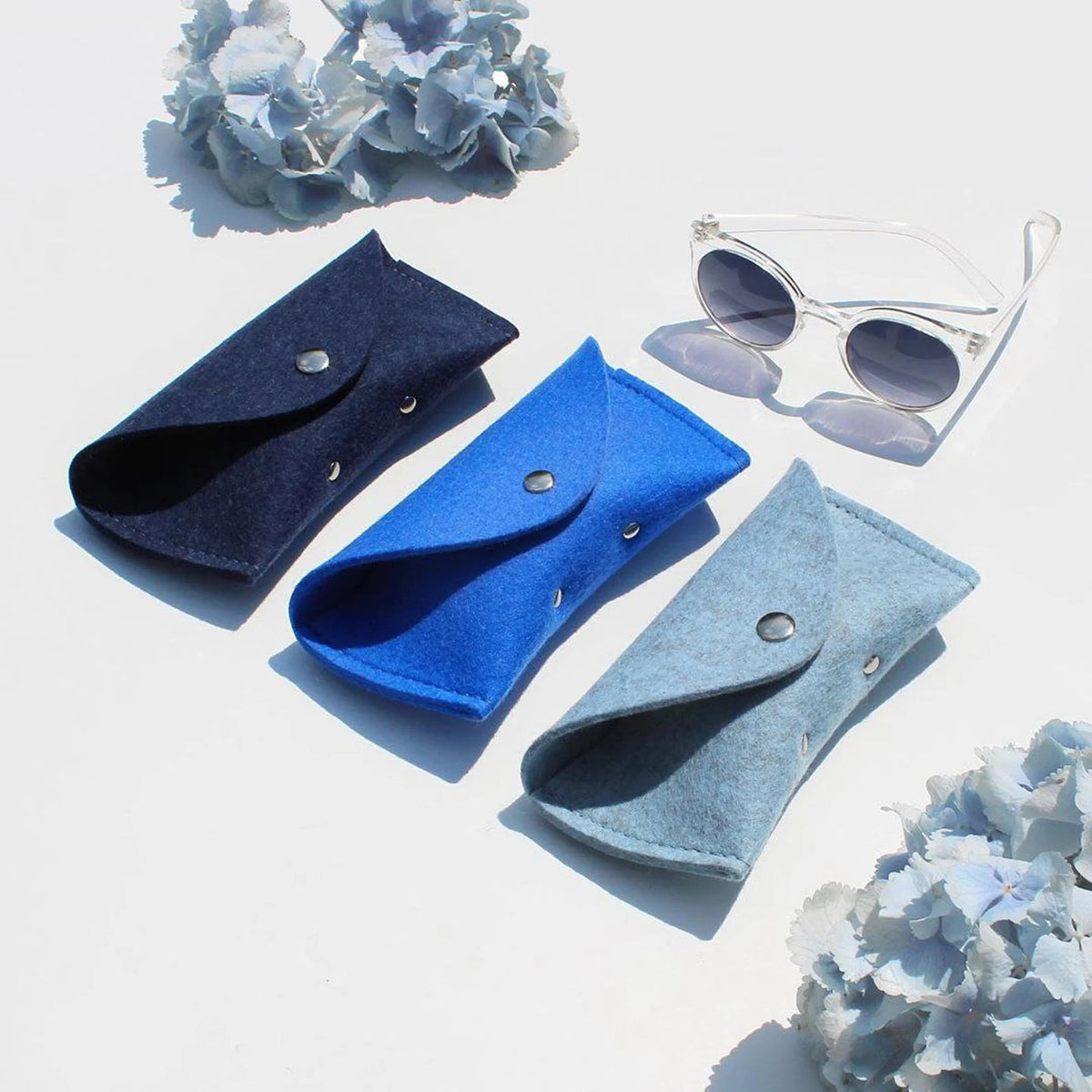 Three WOOLBERRY WOOL FELT GLASSES CASES in navy, bright, and light blue with magnetic closures are arranged on a white surface near clear sunglasses, framed by white-blue hydrangeas.