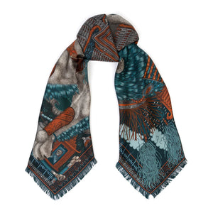 The SABINA SAVAGE - THE SNOW LION SILK TWILL SCARF 135CM by SABINA SAVAGE ENGLAND is a luxurious silk scarf featuring an intricate design in rich browns, blues, and oranges. The pattern, inspired by Tibetan thangkas, includes ornate architectural elements, flowing red hair, and winged figures, all accented by fine detailing and a fringed edge.