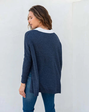 A person with wavy brown hair stands sideways against a plain white background, wearing a light-colored, loose-fitting women's sweater and blue jeans. They glance towards the camera with a neutral expression, showcasing the sporty and stylish appeal of the MERSEA - CATALINA POLO SWEATER by MER SEA.