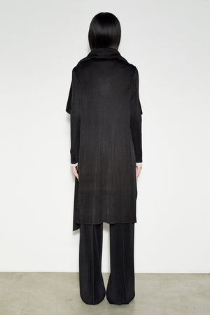 Against a plain white background, a person models the MEIMEIJ - JAPANESE PLEATED LONG JACKET. This black coat exudes Japanese minimalist elegance, paired seamlessly with wide black pants and shoes. The drape front design enhances the depth of this monochromatic ensemble.