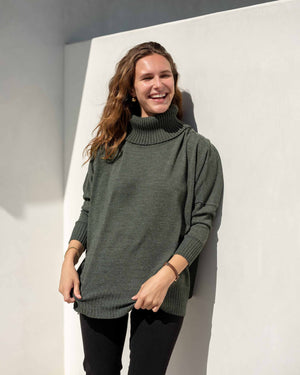 A woman with wavy hair is smiling and looking to the right, wearing a cozy green MERSEA - CATALINA TURTLENECK SWEATER and black pants. She stands against a white wall, with sunlight casting a shadow behind her.