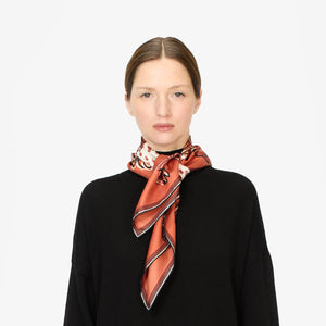 A person with long hair neatly tied back is wearing a sleek, black top and a stylish scarf made in Italy. The INOUI EDITIONS - WESTERN SQUARE SCARF by INOUI, crafted from 100% silk and showcasing rust, cream, and black colors, is elegantly tied around their neck. The plain white background highlights its sophisticated minimalist style, making it perfect for versatile use.