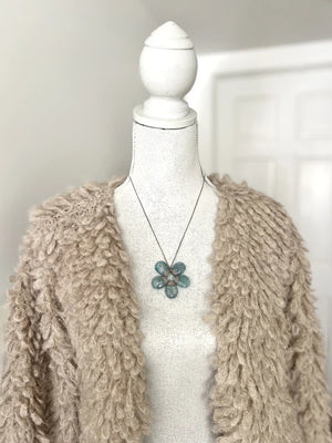 A mannequin in a textured beige cardigan showcases the ANN LIGHTFOOT - BLUE CALCITE FLOWER NECKLACE, highlighted by a striking blue-green pendant. A softly lit white door appears in the background.
