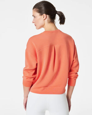 A woman stands confidently against a plain white background, wearing the SPANX Aireessentials Crew Neck Pullover and matching sweatpants made from luxurious comfort spacer fabric. She has her hair pulled back and her left hand is raised slightly. She smiles gently with her lips closed.