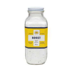 The BAUDELAIRE - BOOST BATH SELTZER 18OZ comes in a clear glass bottle with a yellow label, showcasing white bath salts, essential oils, and visible small herbs inside. The label provides ingredients and usage instructions. The bottle features a screw-on metal cap and is filled nearly to the top.