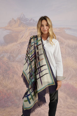 A woman in a blue shirt and jeans stands holding the STORIATIPIC - JULIE WOOL SCARF, a luxury plaid accessory in earthy tones. Behind her, mountains, beach, and grassy dunes create a scenic backdrop.