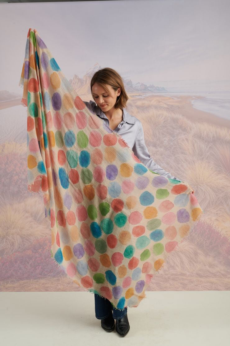 A woman with blonde hair displays the STORIATIPIC - BETTY WOOL SCARF, a large rectangular wool accessory with earth-toned polka dots, set against a scenic mountain landscape.