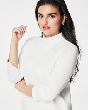 A person with long, dark hair smiles and poses with one hand in their pocket. They are wearing the SPANX - AIRESSENTIALS MOCK NECK TOP made from super-soft fabric, paired with matching dark green leggings against a light background.