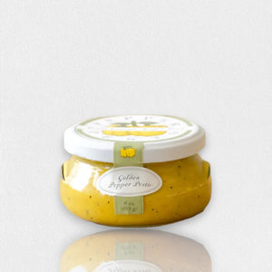 A small, transparent jar of "BELLA CUCINA - GOLDEN PEPPER PESTO" with a white lid. The jar contains a thick, yellow-colored sauce and features a light green label on the front displaying the product name and weight (6 oz. / 169 g). This vegan, small-batch delicacy from BELLA CUCINA is placed on a clean white surface.