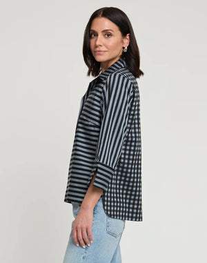 A woman with shoulder-length dark hair wears the HINSON WU - AILEEN 3/4 SLEEVE STRIPE GINGHAM COMBO SHIRT, a black and beige striped blouse featuring a small collar and a partial zip at the front. She pairs it with black pants and stands against a plain white background, one hand in her pocket.