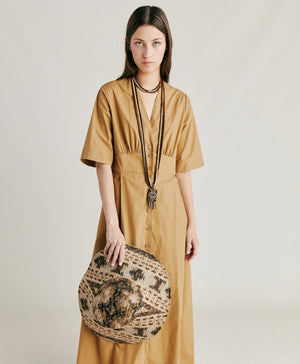 A woman stands in profile wearing the MOMONI - MARTINELLA DRESS, a long, tan cotton poplin dress from MOMONI with short sleeves and a flared skirt. She pairs it with a wide-brimmed, patterned hat, brown sandals, and a necklace. She faces left against a plain, light background.