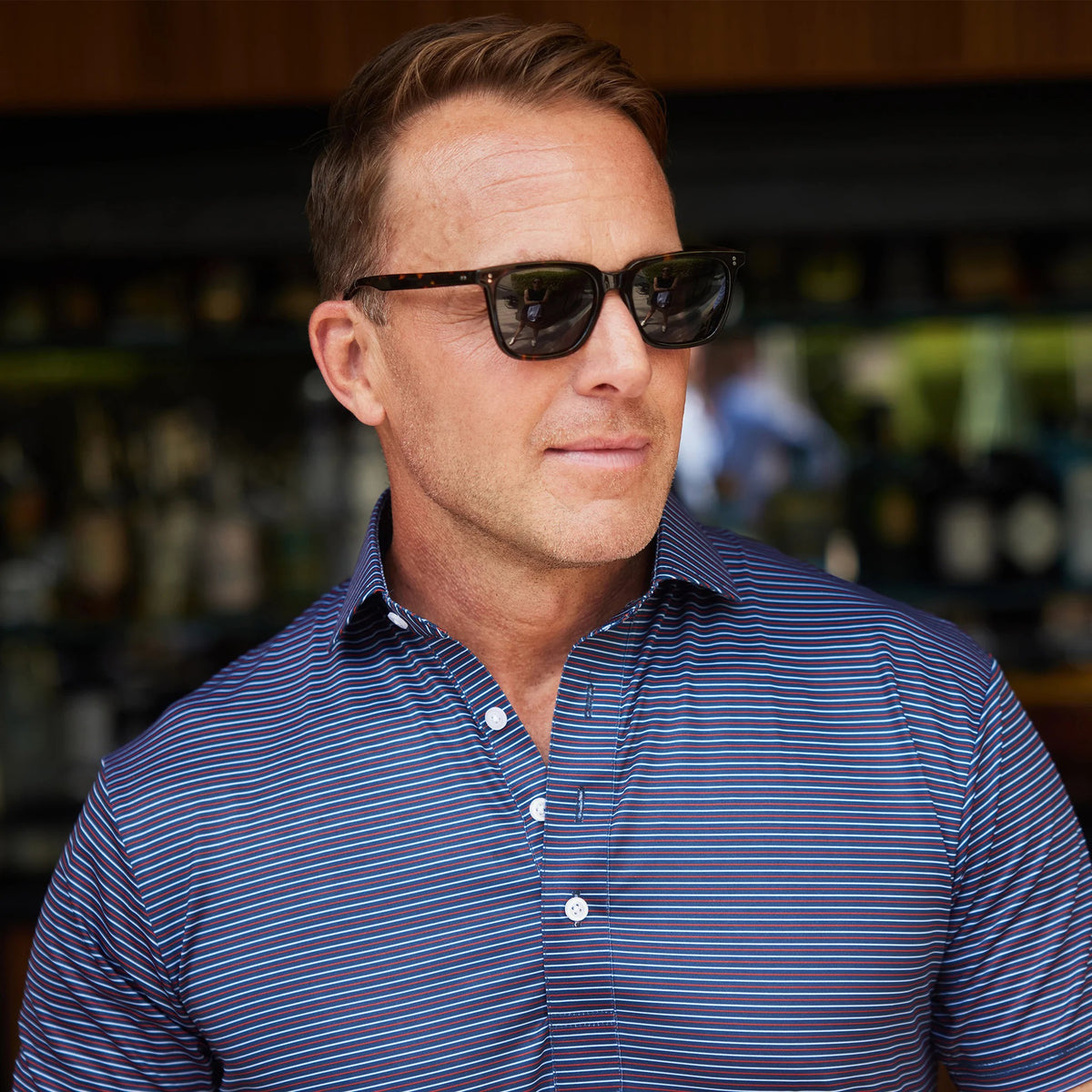 A man wears the COLLARS AND CO - FEATHERWEIGHT FLEX POLO IN LIBERTY NAVY, standing indoors with sunglasses in one hand and a watch on his wrist. The background is blurred, showing shelves of bottles and a dark wooden wall.