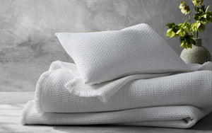 A neatly folded FEARRINGTON LIFESTYLE BEDDING COLLECTION - MATOUK SELAH KING COVERLET by MATOUK, made of 100% cotton, lies on a light gray surface with a matching white textured pillow resting on top. A vase containing green flowers is placed on the right side against a gray, concrete-textured background.