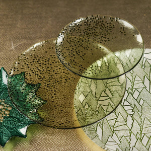 The DOTTED GLASS PLATE 11" by ZODAX is a round ceramic plate with an 11-inch diameter and a green glazed finish. Its surface displays a pattern of small, raised dots that are concentrated on the right side and gradually spread towards the center. The edges of this meticulously crafted plate are slightly uneven.