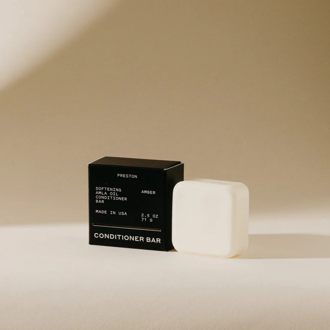 A white conditioner bar rests next to its black packaging on a beige background. The packaging displays &quot;PRESTON,&quot; &quot;Softening Argan Oil Conditioner Bar with Indian Amla Oil,&quot; &quot;Amber,&quot; and &quot;Made in USA,&quot; along with the weight &quot;2.5 OZ&quot; and &quot;71 G.&quot; This plastic-free PRESTON - CONDITIONER BAR nourishes hair for a softer feel.