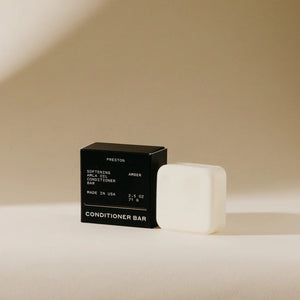 A white conditioner bar rests next to its black packaging on a beige background. The packaging displays "PRESTON," "Softening Argan Oil Conditioner Bar with Indian Amla Oil," "Amber," and "Made in USA," along with the weight "2.5 OZ" and "71 G." This plastic-free PRESTON - CONDITIONER BAR nourishes hair for a softer feel.