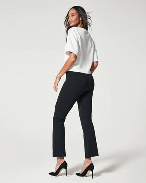 A woman stands confidently against a plain background, smiling broadly. She is wearing a white, short-sleeve top and black SPANX - PERFECT PANT KICK FLARE crafted from smoothing premium ponte fabric and paired with black high heels. Her hands rest on her hips, and her long, dark hair hangs loosely.