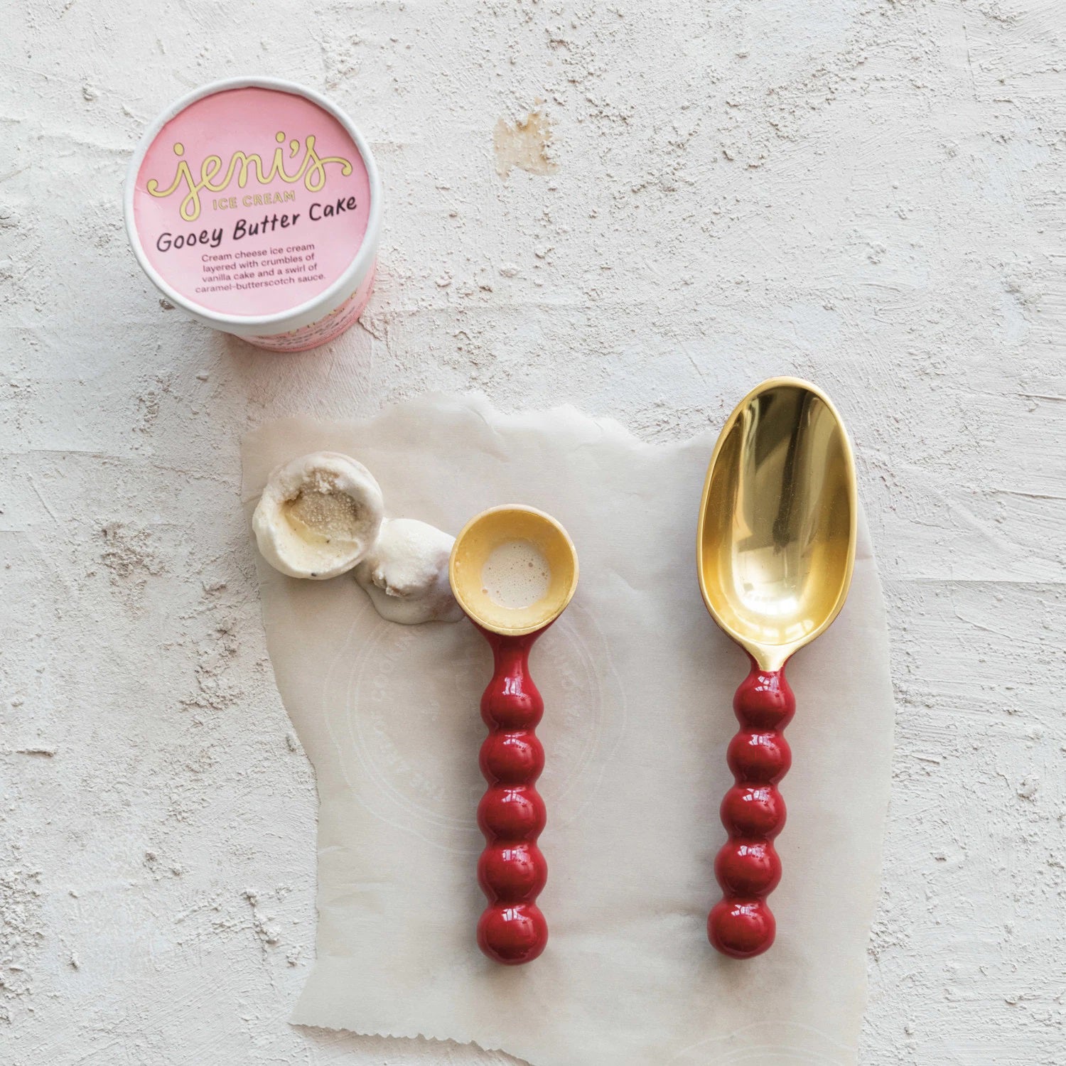 A tub of Jeni's Gooey Butter Cake ice cream sits open on a textured surface. Beside it, two ENAMELED ALUMINUM SCOOPS by CREATIVE COOP, featuring red handles—one small and one large—hold delicious portions of the treat, ensuring a delightful experience that's both stylish and food safe.