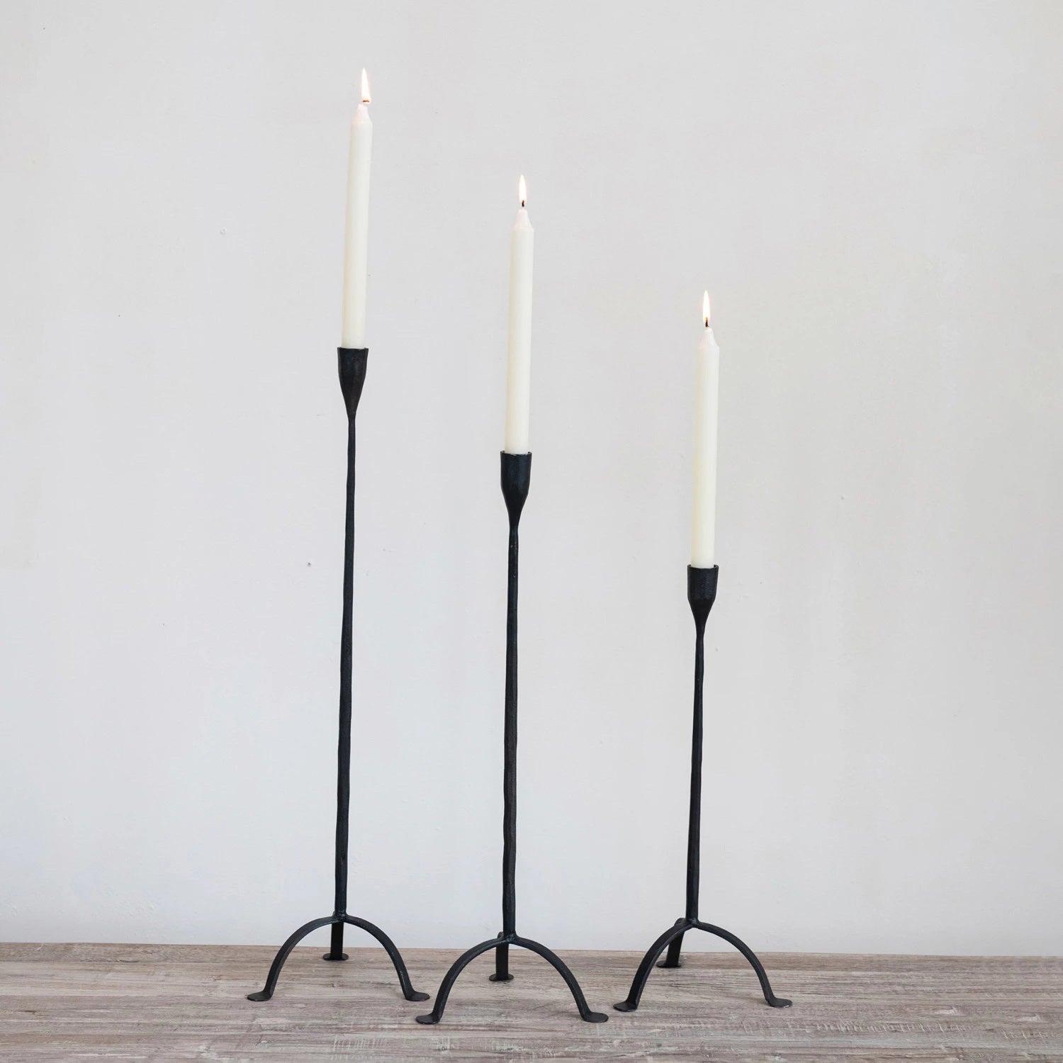 Three Creative Coop HAND FORGED CAST IRON FOOTED TAPER HOLDERS of different heights stand on a wooden surface, each supporting a lit white candle with gently flickering flames, set against a plain, light-colored wall.