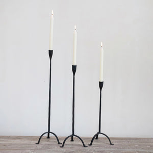 Three Creative Coop HAND FORGED CAST IRON FOOTED TAPER HOLDERS of different heights stand on a wooden surface, each supporting a lit white candle with gently flickering flames, set against a plain, light-colored wall.