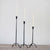 Three Creative Coop HAND FORGED CAST IRON FOOTED TAPER HOLDERS of different heights stand on a wooden surface, each supporting a lit white candle with gently flickering flames, set against a plain, light-colored wall.