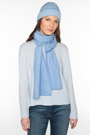 A person wearing a light blue 100% cashmere sweater, a matching light blue beanie, and the KINROSS CASHMERE - PLATED RIB CASHMERE SCARF by KINROSS stands against a plain white background. They have long hair and are looking directly at the camera with a neutral expression.