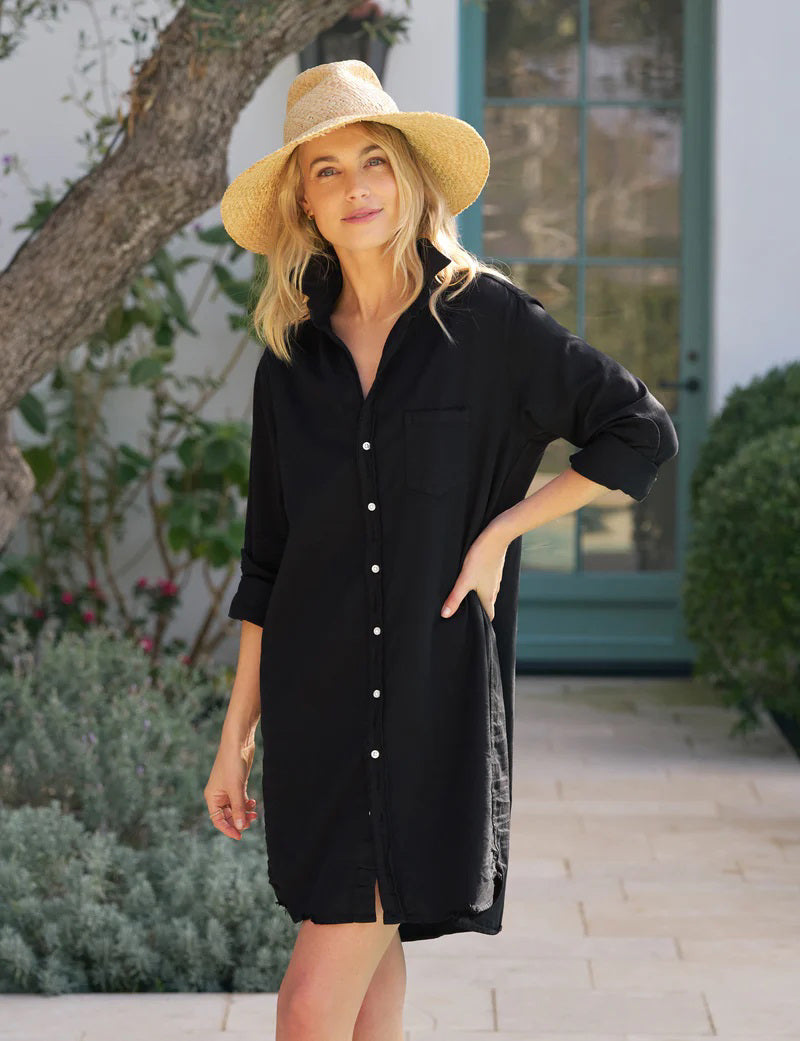 A woman in the FRANK &amp; EILEEN Mary Classic Shirtdress in Famous Denim Black stands outdoors on a stone path, smiling with one hand on her hip. She wears a wide-brimmed straw hat; green plants and a large window are in the background.