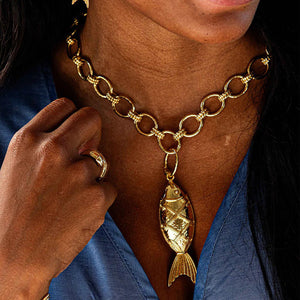 A person wearing the CAPUCINE DE WULF - POISSON PENDANT, featuring large interlocking rings of 18K gold over brass and a golden fish pendant, along with a matching gold ring. The person is wearing a blue top and their hand is touching the good luck talisman.