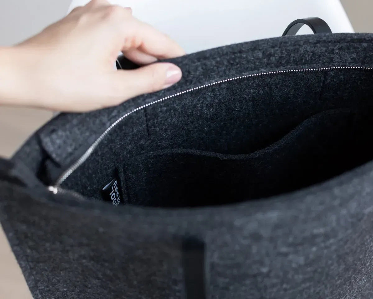 A person in a beige trench coat holds the WOOLBERRY WOOL FELT LONG TOTE BAG with black handles, standing against a plain, light-colored background.