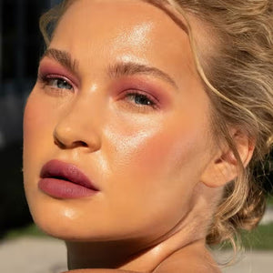 A close-up of a person with curly dark hair, a serious expression, and warm makeup featuring the universally flattering, weightless JASON WU - HOT FLUFF LIP/EYE/CHEEK by JASON WU BEAUTY on their face. They are looking directly at the camera with a neutral background.