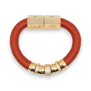 The HOLST + LEE - LUXE CLASSIC BRACELET by HOLST & LEE features an elegant orange cord with gold-plated accents, including a rectangular gold clasp adorned with several gold and diamond-encrusted beads near the clasp.