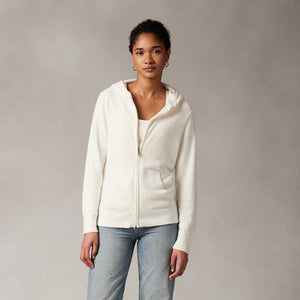 A person with brown skin and dark hair is wearing a white KASHWERE - RELAXED HOODIE made from Kash-ease Yarn. They are looking down while gently holding the edge of the hood with one hand against a simple, neutral-colored background.