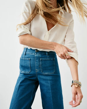 A person with long hair, partially covering their face, is wearing a white button-up shirt with the sleeves slightly rolled up. They are also sporting the SPANX EveryWear Cropped Wide Leg Jeans with patch pockets, along with a watch and bracelet on their wrist.