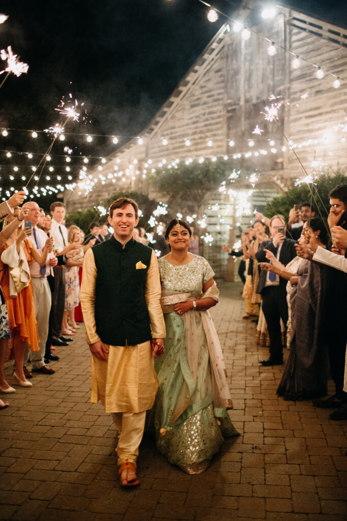 A couple in traditional Indian attire walks hand-in-hand under string lights, surrounded by guests holding sparklers, outside a rustic barn at night during a festive celebration.