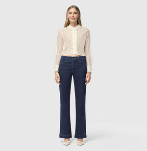 A person with light brown hair stands against a plain background, wearing a sheer, ruffled, long-sleeved white blouse and MAC JEANS - DREAM WIDE AUTHENTIC by MAC in dark blue. They're paired with comfortable white shoes.