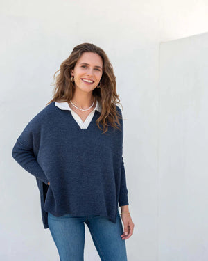 A person with wavy brown hair stands sideways against a plain white background, wearing a light-colored, loose-fitting women's sweater and blue jeans. They glance towards the camera with a neutral expression, showcasing the sporty and stylish appeal of the MERSEA - CATALINA POLO SWEATER by MER SEA.