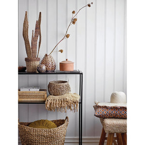 A neatly arranged shelf and stool are adorned with CREATIVE COOP SEAGRASS BASKETS WITH HANDLES, featuring circular handles. These baskets, along with a blanket, a hat, a plant, and decorative pottery against a white paneled wall, create a warm, rustic vibe with natural tones and textures.