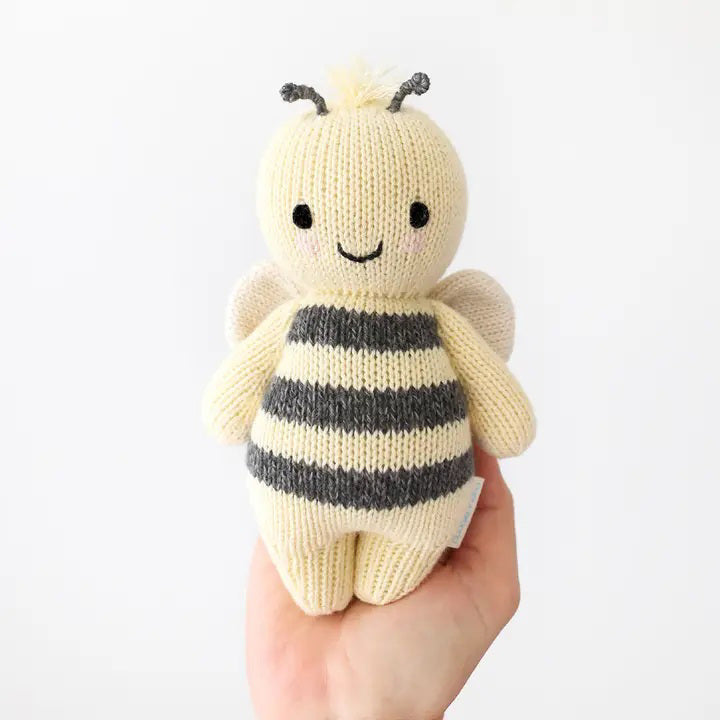 A baby lies on a cozy white blanket, raising both arms. The baby is dressed in a peach outfit and a headband with three pom-poms. Next to the baby is the CUDDLE + KIND - HANDMADE BABY BEE by CUDDLE &amp; KIND, a fair trade, hand-knit toy with black and white stripes—a perfect addition to any collection of baby shower gifts.