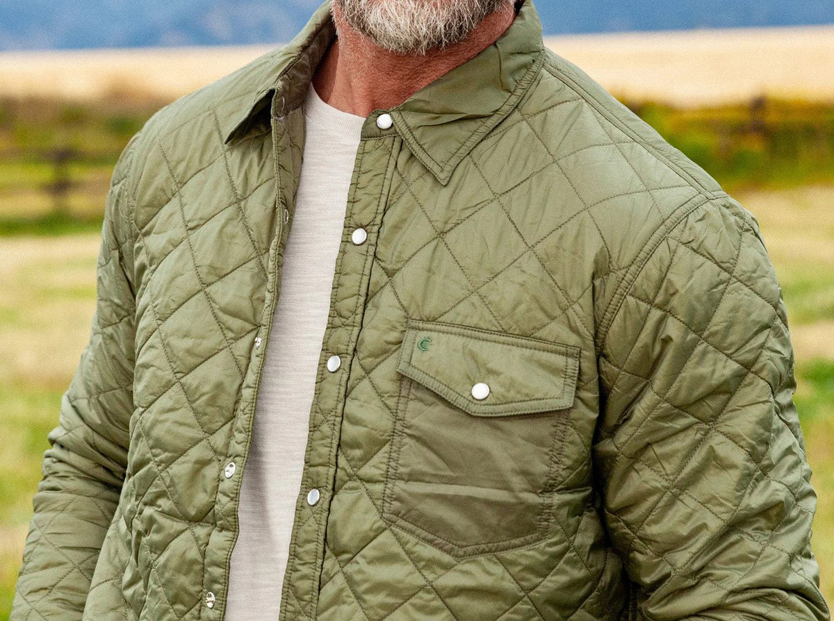 A smiling bearded man stands outdoors in a grassy field, wearing the CRIQUET Quilted Shacket by CRIQUET, paired with jeans and holding a canned drink. Mountains and a wooden fence can be seen in the background.