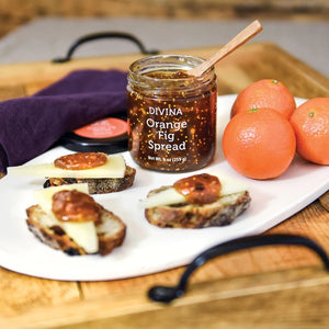 A glass jar of DIVINA - ORANGE & FIG SPREAD by GOURMET FOOD SOLUTIONS, featuring a black lid, is displayed. The label indicates a net weight of 9 ounces (255 grams). Reminiscent of traditional fig jam, this product is certified non-GMO and vegan, as indicated by the icons on the bottom right of the label.