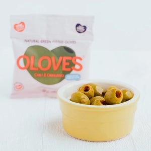 A small, light pink packet of POSHI brand olives named OLOVES - CHILI & OREGANO. The package features the text "Natural Green Pitted Olives" and "Chili & Oregano" with a green olive illustration. Labels indicate it's a low-calorie, gluten-free vegan snack with a new Greek recipe. Net weight: 30g / 1.1 oz.