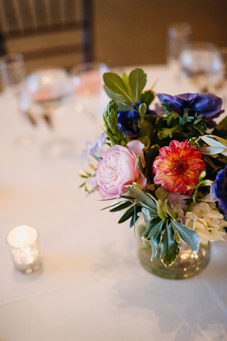 A small glass vase with pink, red, and purple flowers and green leaves sits on a white tablecloth near a lit votive candle, with blurred tableware in the background.
