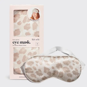 A LEOPARD SATIN SLEEP MASK from KITSCH, featuring a chic beige and white leopard print design, is displayed in front of its packaging. The packaging includes a cutout showcasing the sleep mask alongside an image of a woman wearing it. Text on the packaging reads: "The KITSCH Satin Eye Masks for deeper sleep," highlighting this luxurious satin product from a female-owned company.