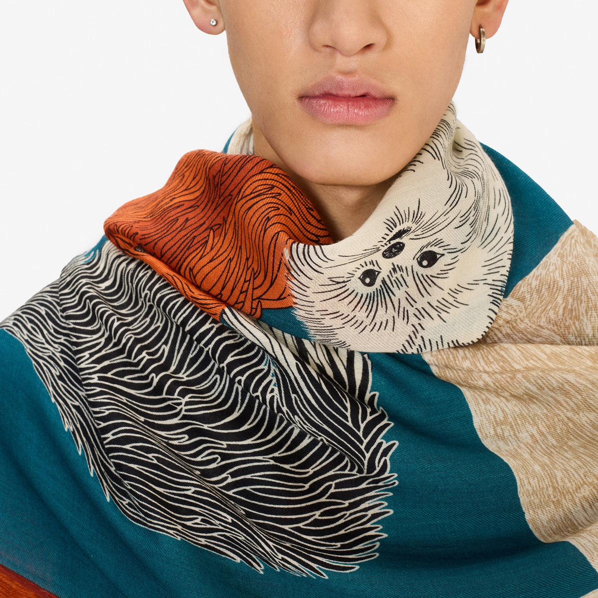 A woman with short blonde hair wears a dark jacket and the INOUI EDITIONS TOUTOU Dogs wool scarf in duck blue from INOUI, featuring playful animal illustrations in orange, teal, and cream, standing against a plain white background.