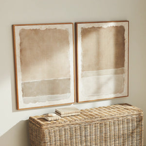 Two NAPA HOME AND GARDEN - COMPOSITION IN TAUPE GICLEES with wooden frames hang on the wall. Below them is a woven rattan console table adorned with books and stones, creating a timeless look in the gently sunlit space.