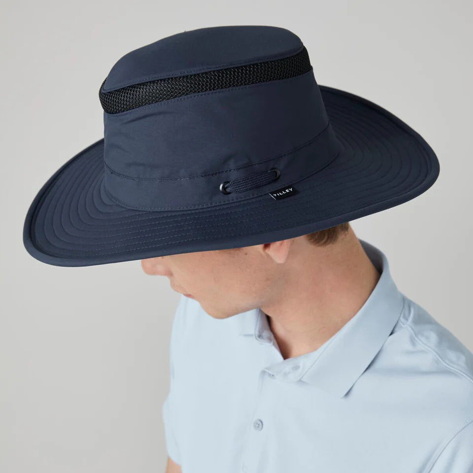 A person wearing the TILLEY - AIRFLO BROAD BRIM LTM6 hat with a light blue collared shirt, looking downward against a plain, light background.