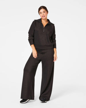 A person is wearing the SPANX AIRESSENTIALS WIDE LEG SHIMMER PANT in black along with a zip-up top. They stand casually with their hair tied back, sporting black and white sneakers, all set against a plain white background.