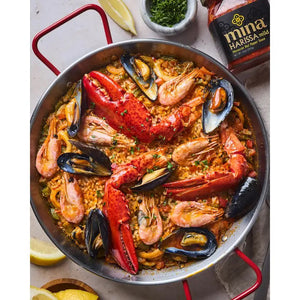 A seafood paella in a large round pan, filled with rice, shrimp, mussels, clams, and lobster claws, garnished with herbs. A jar of MINA - HARISSA ORIGINAL SPICY from the brand MINA adds a spicy harissa kick to the dish. Lemon wedges and a small bowl of chopped herbs are placed beside the pan on a light wooden surface.