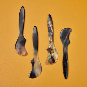 Four uniquely shaped and polished HORN SPREADERS by BE HOME, INC., handmade in Vietnam, are displayed against a warm mustard yellow background. Each spreader exhibits a distinct wavy design and a blend of dark and light natural hues.