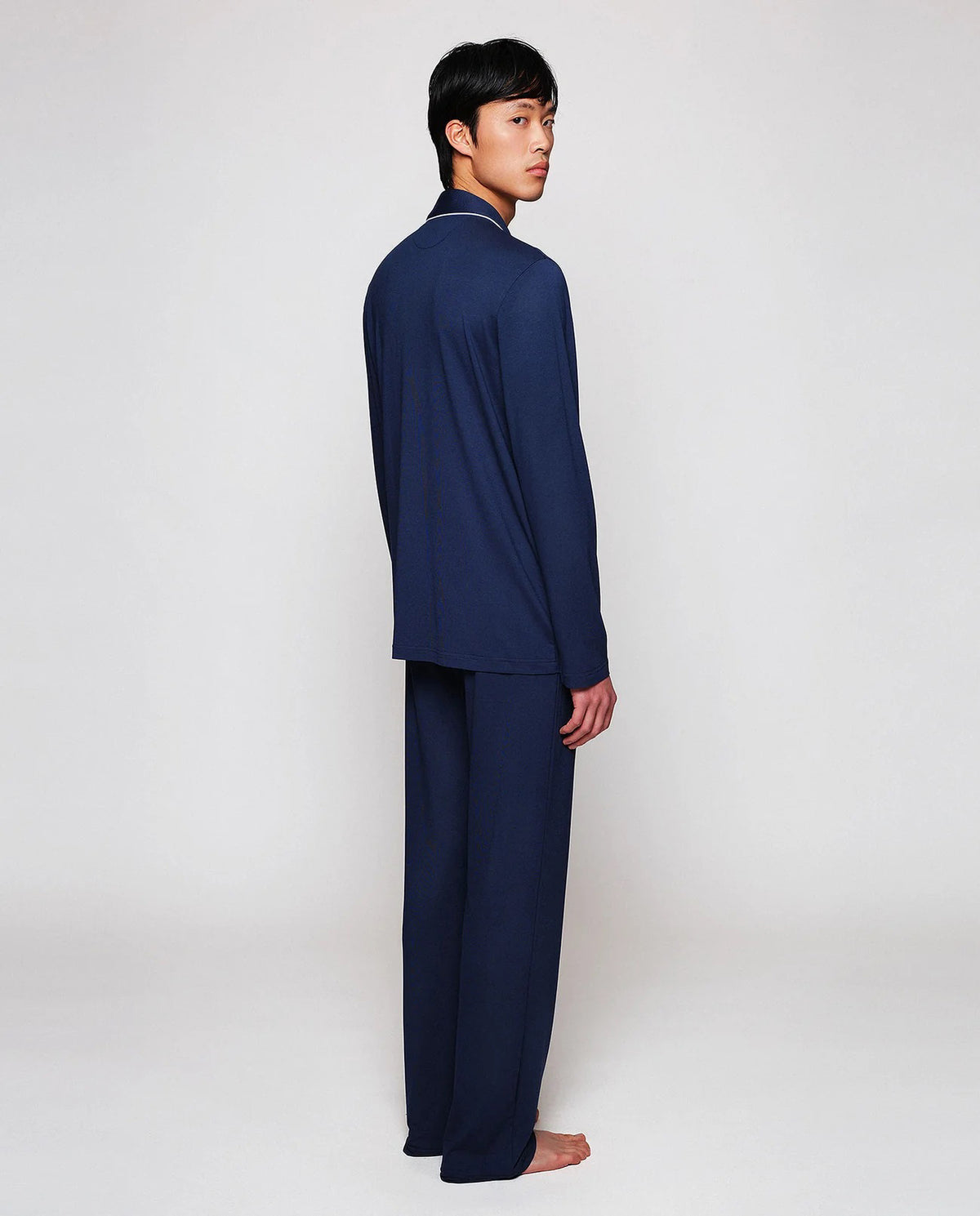 A person with short dark hair stands barefoot on a white background, wearing the MIRTO 1956 - NAVY MODAL PAJAMAS: a breathable matching set with white piping and buttons by MIRTO 1956.